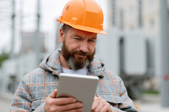 Bearded man wears orange hard hat. He holds tablet, focused on screen. Industrial background suggests field work. Concentrated, he checks data or plans. Useful for construction, engineering