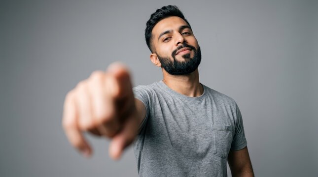 Young man with a beard pointing toward the lens in a studio with a gray background