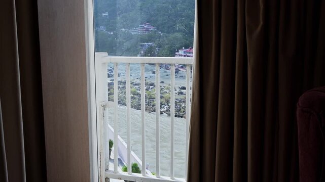 Curtains frame an open balcony view of the Ganga River, white railings and wooded hills in Rishikesh.