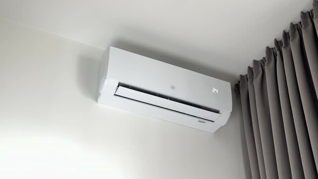 Hand holding remote controller to turn on a white split system conditioner. Wireless device activates the home climate technology to open its horizontal vent flap. Wall mounted modern appliance