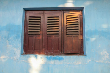 Blue wall frames aged wooden shutters. Sunlight dances on textured surface, casting soft shadows. Shutters show wear, hinting at history and quiet stories. Ideal for rustic, vintage, or coastal themes © Restu