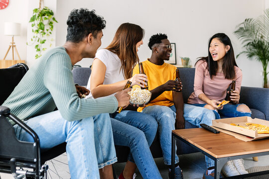 Group of diverse friends sharing popcorn and pizza together at home. Young multiracial people laughing during a casual movie day. Friendship and community concept.