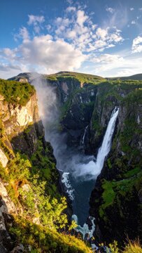 Majestic waterfall cascades down a lush green cliffside under a cloudy blue sky.