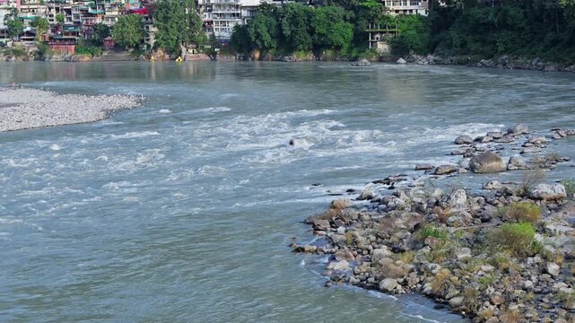 Whitewater ripples over rocks in the Ganga River below a colorful riverside settlement in Rishikesh.