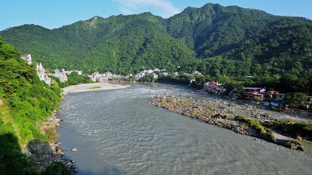 A rocky gravel bar divides the flowing Ganga River below hillside buildings in Rishikesh.