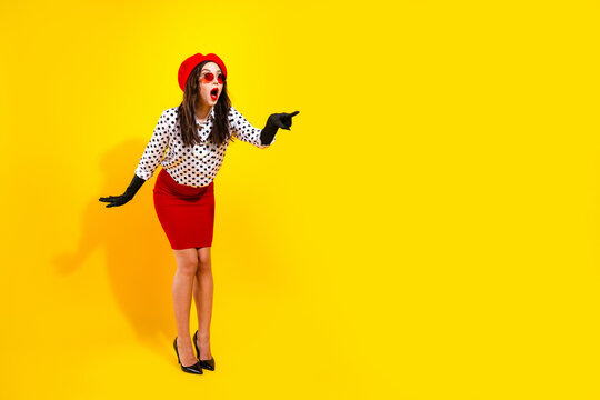 Stylish surprised woman in red skirt and beret pointing against vibrant yellow background, vintage polka dot blouse and sunglasses fashion portrait conveying excitement and playful retro attitude