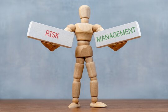 Wooden doll standing on a wooden desk holding two white blocks reading RISK and MANAGEMENT. Corporate governance, strategic balance, business decision-making concept.