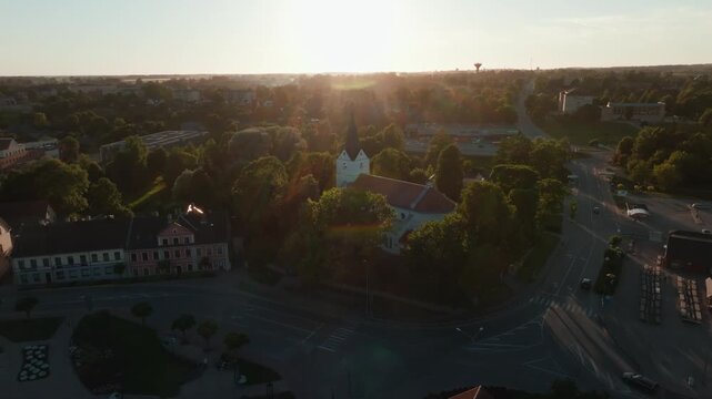 Saldus town square to church steeple at sunset from drone, Latvia. 4K aerial footage crossing historic paved plaza, red tiled roofs and trees toward white lutheran church.