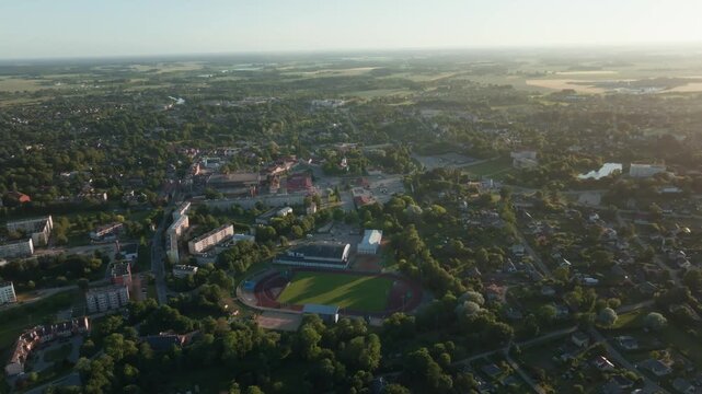 Small town with stadium and apartment blocks from high drone at sunset. 4K aerial footage gliding high over football pitch, running track, apartment buildings, private houses, roads.