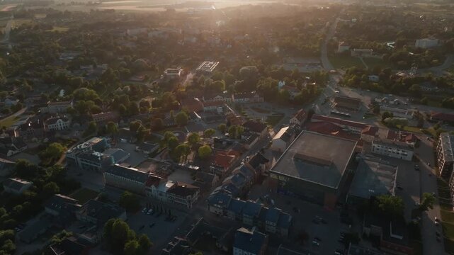Town center rooftops from drone flying toward sunset haze. 4K aerial footage passing large flat roof building, historic houses, red tiled roofs, apartment blocks, streets.