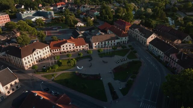 Orbiting drone shot of Saldus town square in evening light, Latvia. 4K aerial footage circling historic paved plaza with lawns, flower beds, trimmed trees, sculpture, old buildings.