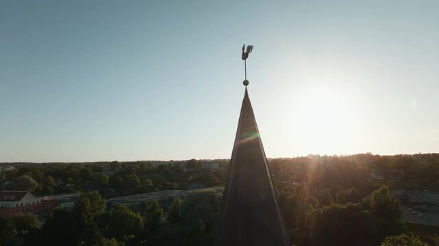 Rooster weathervane on Saldus church steeple from drone at sunset, Latvia. 4K aerial footage moving from spire silhouette against sun to golden copper cockerel above historic town square.