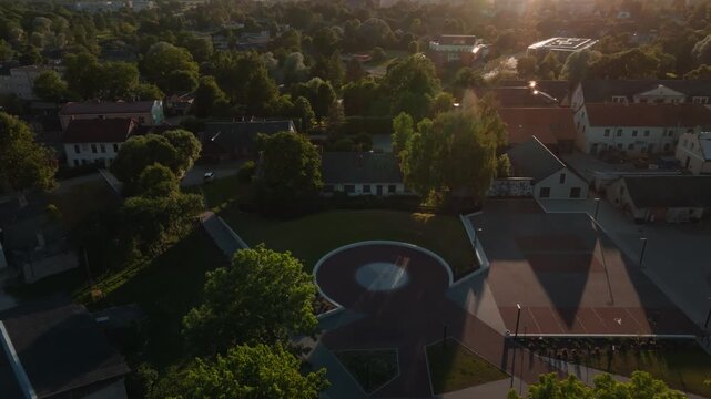 Circular plaza in Saldus old town from retreating drone at sunset, Latvia. 4K aerial footage retreating from renovated round square with green lawn over historic houses, red tiled roofs.