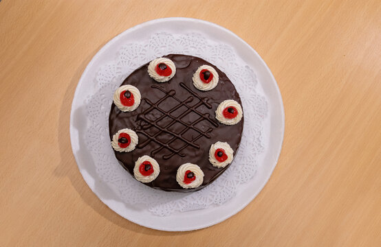 Vista superior de pastel de chocolate glaseado con crema y cerezas sobre plato blanco con blonda, en mesa de madera clara.