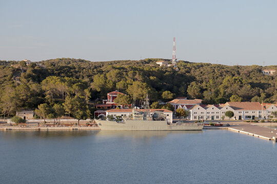Buque militar patrullero amarrado en el muelle de la base naval de Mah&oacute;n, rodeado de colinas boscosas y arquitectura mar&iacute;tima tradicional en Menorca.