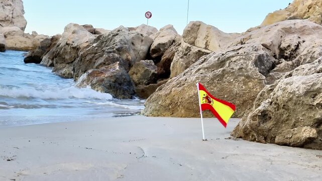 Spanish flag positioned on sandy beach near rocky coastline, with gentle waves lapping at the shore and a no swimming sign visible in the background, creating a serene coastal scene