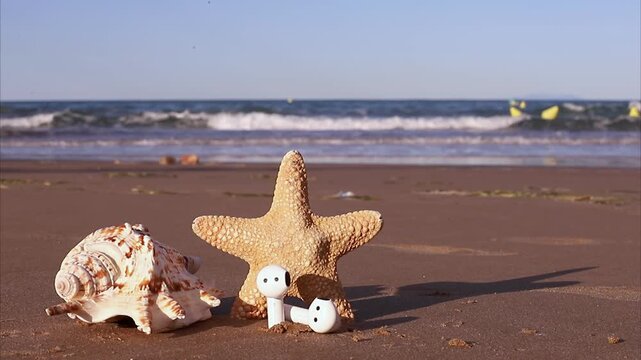 Shell and starfish arrangement on sandy beach with wireless earbuds positioned near the ocean waves, showcasing a serene coastal scene under clear blue skies and gentle sunlight