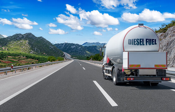 A white fuel tanker truck drives along an empty highway surrounded by scenic mountain landscapes under a sunny blue sky.