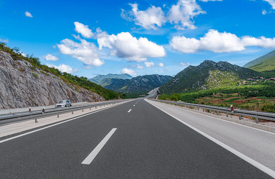 An open asphalt highway stretches through a scenic mountain landscape under a bright blue sky with fluffy white clouds.