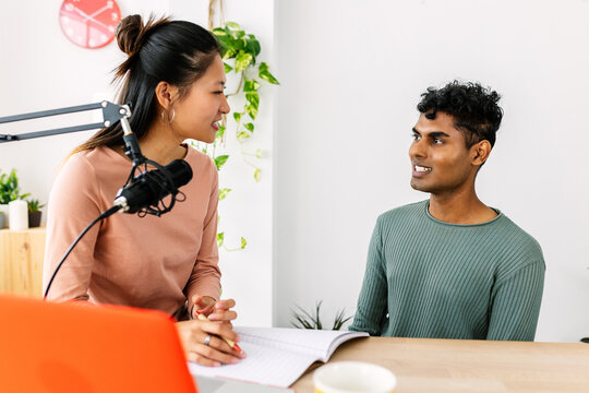 Young woman interviewing her Indian friend with a microphone for a podcast at home, both smiling. Diverse multiracial people creating content together. Friendship and technology concept.