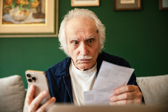 Shocked senior man checking bills on phone at home