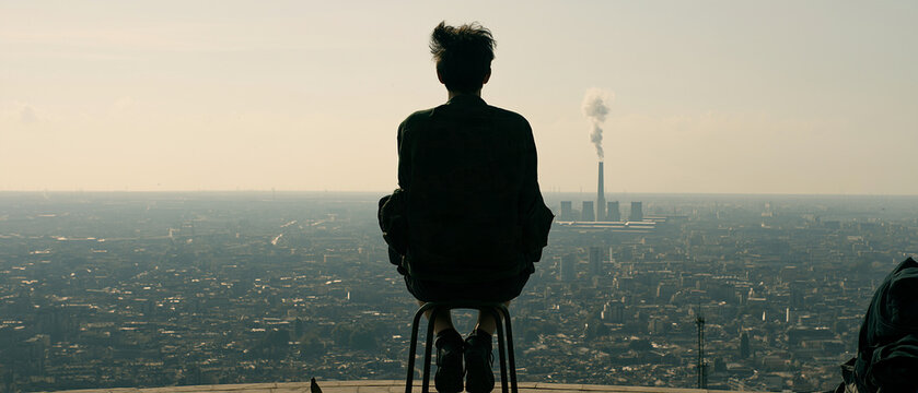 Silhouette Contemplation: A solitary figure sits in quiet contemplation, silhouetted against a sprawling urban landscape and a distant industrial horizon.