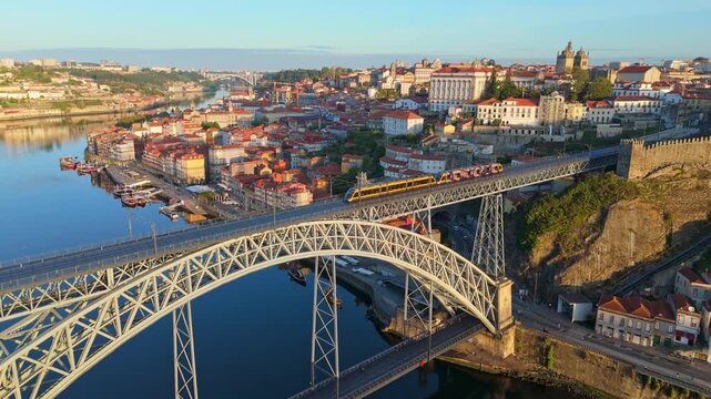 Drone view of Dom Luis I Bridge over Douro River in Porto, Portugal, with upper deck details, including a tram on the bridge and rabelo boats by the quay at sunrise. Orbit parallax shot