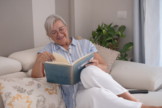 Senior woman relaxing on a comfortable sofa at home, enjoying a book and smiling, finding joy in her hobby and leisure time during retirement