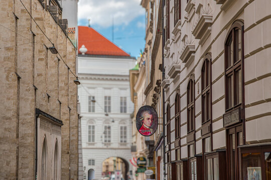 A narrow street in Vienna's Old Town, Austria, near the Hofburg / Augustinerstra&szlig;e area, distinguished by buildings with classical architecture and a hanging sign featuring a portrait of W. A. Mozart.