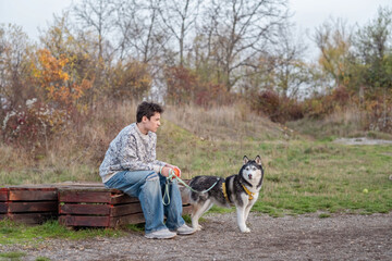 Husky reaches to teenager sitting on wooden bench in city park, friendship built through daily quiet hours, scene for pet brand. Wide shot. Concept of friendship between teenager and husky © iwavephoto