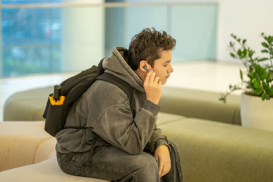 Teenage boy adjusts wireless earphone sitting on bench near plants, sitting down after long walking hour, moment parents rarely see. Wide shot. Concept of sitting pause during shopping day