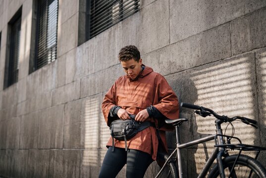 A young urban cyclist wearing a waterproof rain poncho adjusts their waist bag while standing next to a bicycle by a stone wall