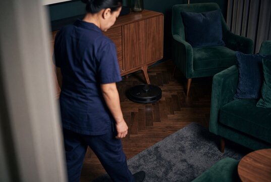 Housekeeper Watching a Modern Robotic Vacuum Cleaner Cleaning Hardwood Floor in a Stylish Living Room