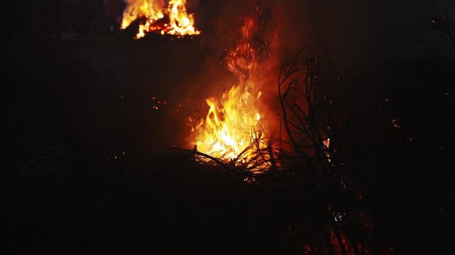Intense flames from a large bonfire devouring dry brush at night. Bright orange and yellow fire rises with thick smoke and glowing sparks