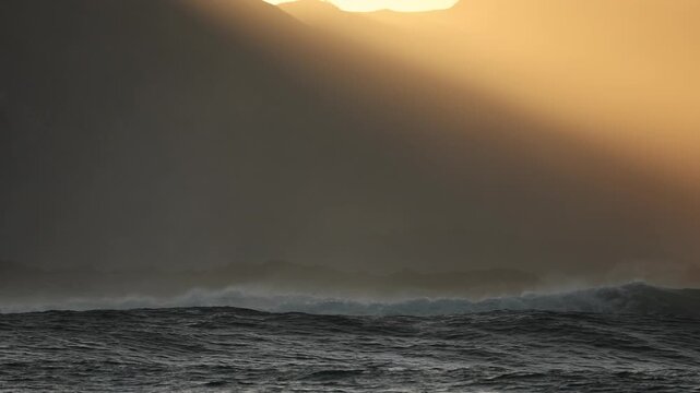 A breathtaking golden sunset illuminates the ocean along the coast of South Africa, casting warm reflections across the water. A large dark wave rises dramatically in the foreground, creating a