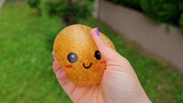 golden kawaii squishy beaded balls with cute smiley face held in hand, sparkly orbeez textured toys squeezed outdoors on green background