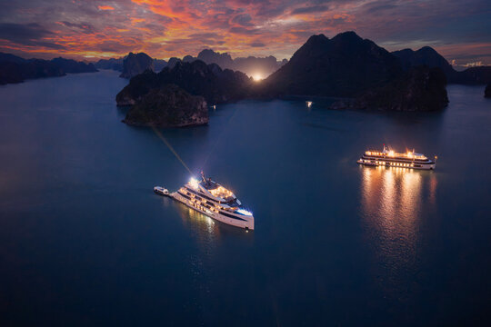 Aerial view of illuminated cruise ships anchored in Ha Long Bay, Vietnam, at dusk with dramatic sunset clouds and limestone karsts