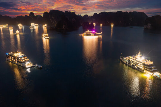 Aerial view of illuminated cruise ships anchored in Ha Long Bay, Vietnam, at dusk with dramatic sunset clouds and limestone karsts