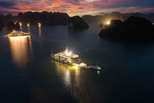 Aerial view of illuminated cruise ships anchored in Ha Long Bay, Vietnam, at dusk with dramatic sunset clouds and limestone karsts
