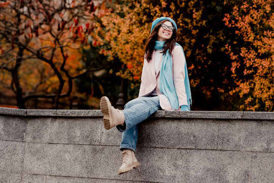 Smiling young woman sitting on a stone wall in autumn wearing a blue scarf and beret enjoying fall outdoors