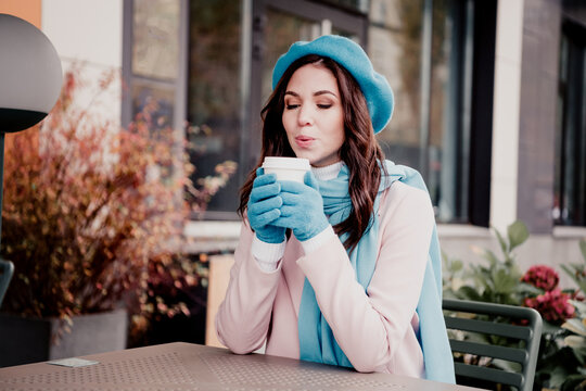 Young woman in a blue beret and scarf enjoying a hot coffee outdoors at a caf&eacute; on a chilly autumn day