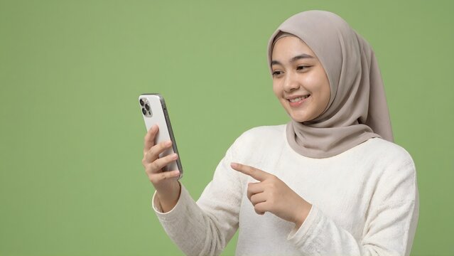 Smiling Muslim woman in hijab using and pointing at smartphone