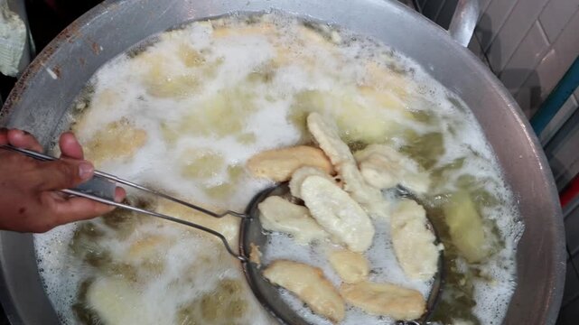 Close up of vendor's hand scooping golden banana fritters from bubbling transparent oil in a pan until empty