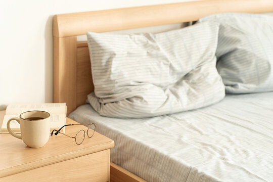 Cozy, leisurely morning ritual in bright bedroom: cup of coffee, an open book, and glasses on wooden nightstand, soft natural light, and comfortable unmade bed. Slow living and mindful lifestyle