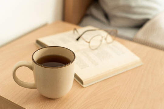Cup of warm tea in mug on light wooden nightstand, an open book and reading glasses create cozy atmosphere for relaxation. Slow living, mindful hobby