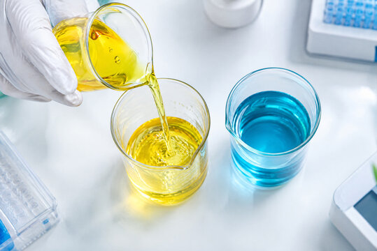 A scientist or technician pours yellow liquid into a glass, with additional glasses containing blue and yellow liquids on a lab bench, illustrating a scientific or chemical experiment setup.