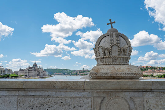 Holy Crown of Hungary sculpture on a bridge parapet framing a distant view of the Hungarian Parliament across the Danube