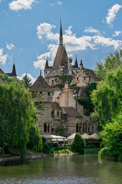 Vajdahunyad Castle towers rising above willow trees and lakeside cafe umbrellas in City Park Budapest