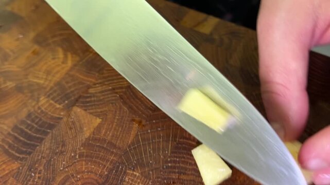A hand holds a pickled cucumber while a kitchen knife cuts it into small pieces on a textured wooden cutting board.
