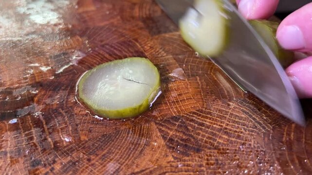 A kitchen knife slices pickled cucumber into thin rounds on a wet wooden cutting board, revealing its translucent flesh and seeds.